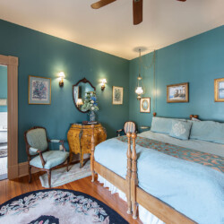 A traditionally styled bedroom with teal walls features a large bed, wooden furniture, and framed artwork. An adjacent bathroom with a clawfoot tub is visible through the open door.