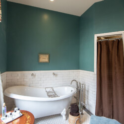 A bathroom with teal walls, white subway tile wainscoting, a freestanding bathtub, and a brown shower curtain. A wooden table with toiletries is beside the window, and a basket with towels sits on the floor.