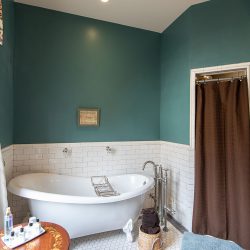 A bathroom with teal walls, white subway tile wainscoting, a freestanding bathtub, and a brown shower curtain. A wooden table with toiletries is beside the window, and a basket with towels sits on the floor.