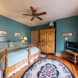 A bedroom with teal walls features a wooden bed, a TV on a dresser, a ceiling fan, an armoire, framed artwork, and two rugs on the wooden floor. A window with floral curtains allows natural light.