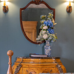 Ornate wooden dresser with a floral arrangement in a blue vase, a book, and a mirror mounted above. Twin wall sconces with shades flank the mirror against a teal wall. Corner of a bed in the foreground.