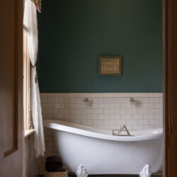 A vintage-style bathroom with a clawfoot bathtub, white subway tiles, green walls, a window with drapes, a wicker basket, and a floral rug on a mosaic-tiled floor.