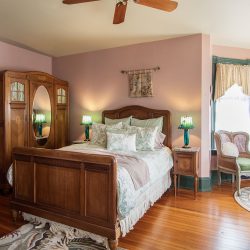 A spacious bedroom with wooden furniture including a large bed, wardrobe, and bedside tables. The room has pink walls, a ceiling fan, green armchairs, and a window with lace curtains.