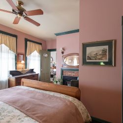 A bedroom with pink walls, a wooden bed, floral bedding, a ceiling fan, windows with draped curtains, and a small TV. An adjacent bathroom with a shower is visible.