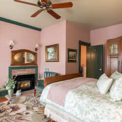 A cozy bedroom featuring a bed with floral-patterned bedding, a brick fireplace, a ceiling fan, green trim, and various pieces of furniture and decor including framed artwork and a large armoire.