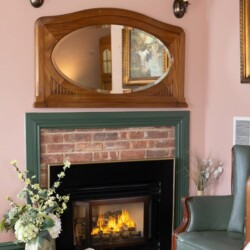 A cozy fireplace with a brick surround and black insert. Above it, there's a large wooden framed mirror. Nearby are a green armchair, a decorative vase with flowers, and wall sconces with lampshades.