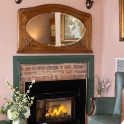 A cozy fireplace with a brick surround and black insert. Above it, there's a large wooden framed mirror. Nearby are a green armchair, a decorative vase with flowers, and wall sconces with lampshades.