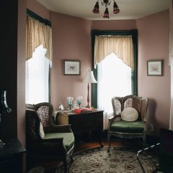 A cozy room with two cushioned chairs, a round patterned rug, a small table with a lamp, and decor framed by two large windows with light curtains.