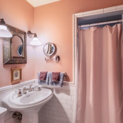 A bathroom with pink walls features a white pedestal sink, wall-mounted mirror, lights, pink shower curtain, and towels on a rod.