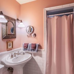 A bathroom with pink walls features a white pedestal sink, wall-mounted mirror, lights, pink shower curtain, and towels on a rod.