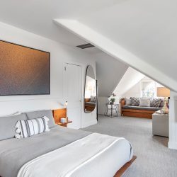 A modern bedroom with a sloped ceiling features a large bed, wall-mounted TV, oval mirror, abstract painting, and a cozy seating area by the window. The room has neutral tones and minimalist decor.