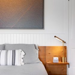 A neatly made bed with grey linens and a striped cushion. A wooden nightstand with a brass lamp and a small box sits beside it. A large abstract painting hangs on the wall above the headboard.