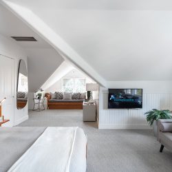 A spacious, modern bedroom with angled ceilings, a mounted TV, and a sitting area by the window. There's a bed, a sofa, wall art, and a wooden nightstand with a lamp. The room is brightly lit.