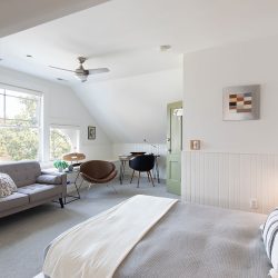 A bright, modern bedroom with a vaulted ceiling, a bed, a sofa, a wooden table with chairs, and an open window letting in natural light. There's also a ceiling fan and a wall-mounted artwork.