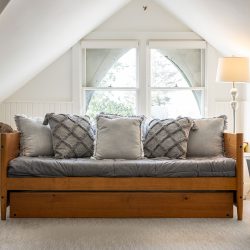 A small, cozy loft with a wooden daybed topped with gray pillows, a table lamp, a houseplant, and a metallic floor lamp. Light filters in through a double window with arched upper panes.