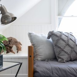 A cozy room with a gray daybed, decorated pillows, a modern metal lamp, a potted plant, and a black alarm clock on a side table. Natural light filters through a window in the background.
