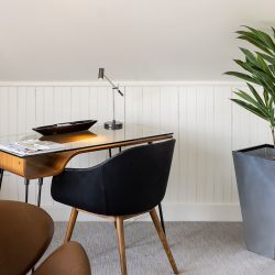 A modern home office space with a glass-top desk, black chair, desk lamp, and magazines. A potted plant stands next to the desk against a white wainscoted wall.