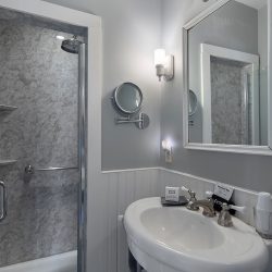 Small bathroom with a shower enclosed by a glass door, a white sink with a mirror above, and a wall-mounted light fixture. There are toiletries on the sink and a magnifying mirror on the wall.