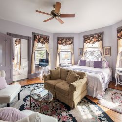 A bright bedroom with a double bed, floral curtains, a ceiling fan, a small round glass coffee table, a beige sofa, and a cushioned armchair. The room features hardwood floors and a floral rug.
