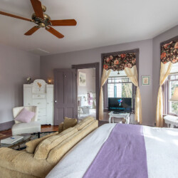 A cozy bedroom with light purple walls, a ceiling fan, floral curtains, a bed, a fireplace, a sofa, a dresser, and a television. Sunlight streams in through a window beside a table with a lamp.