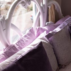 A close-up of a bed with a white ornate headboard, light purple and lavender pillows, and light streaming in through a window. Light purple curtains hang to the side.