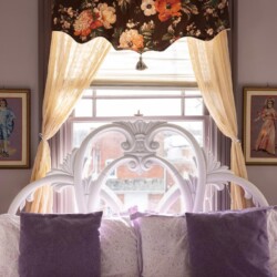 A bed with a white ornate headboard, purple and white bedding, and violet pillows, is positioned in front of a window adorned with floral drapes and sheer curtains, flanked by two paintings.