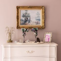 A decorative mantle with a framed painting, wine glasses with floral decorations, a framed pink photo, a pine cone, and various ornaments on a vintage white dresser against a pink wall.