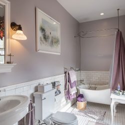 A bathroom with a white sink, a toilet, a clawfoot bathtub with a purple shower curtain, a window with floral curtains, and light purple walls. There are various toiletries and a wicker basket.