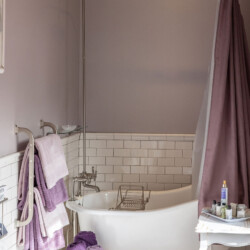 A bathroom with a freestanding bathtub, purple shower curtain, wall-mounted showerhead, white subway tile, and purple towels on a rack. A basket of towels and a side table with toiletries are also visible.