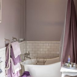 A bathroom with a freestanding bathtub, purple shower curtain, wall-mounted showerhead, white subway tile, and purple towels on a rack. A basket of towels and a side table with toiletries are also visible.