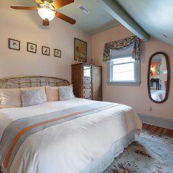 A bedroom with a brass bed frame, white bedding, wooden dresser, oval mirror, table with a TV, and a ceiling fan. The room has floral curtains, a patterned rug, and framed art on the walls.