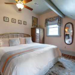 A bedroom with a brass bed frame, white bedding, wooden dresser, oval mirror, table with a TV, and a ceiling fan. The room has floral curtains, a patterned rug, and framed art on the walls.