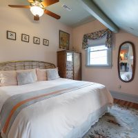 A bedroom with a brass bed frame, white bedding, wooden dresser, oval mirror, table with a TV, and a ceiling fan. The room has floral curtains, a patterned rug, and framed art on the walls.