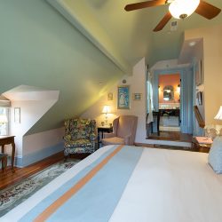 A cozy bedroom with a slanted ceiling, armchair, patterned chair, small table, and bed with light blue and orange bedding. Lamps and wall decor are present, and a bathroom is visible through an open door.