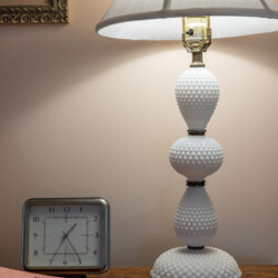 A table lamp with a scalloped base sits on a wooden table alongside a square clock, a pink folder, and a pen.