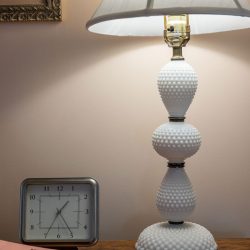 A table lamp with a scalloped base sits on a wooden table alongside a square clock, a pink folder, and a pen.