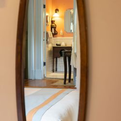 A bedroom mirror reflects a view into a bathroom with orange walls, a lit sconce, a sink, and tiled flooring. The edge of a bed is visible in the foreground.