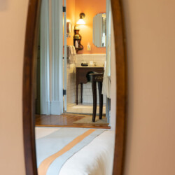A bedroom mirror reflects a view into a bathroom with orange walls, a lit sconce, a sink, and tiled flooring. The edge of a bed is visible in the foreground.