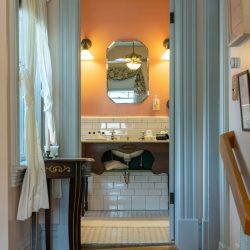 A view through a doorway into a vintage bathroom with orange walls, a wooden sink with tiled backsplash, white and light blue accents, and a mirror illuminated by two wall-mounted lights.