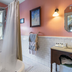 A bathroom with white tile walls, a pink upper wall, a bathtub with a curtain, a vanity sink with toiletries, towels hanging on a rack, a window with curtains, and a mirror with a light above it.
