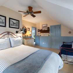 A cozy bedroom with a large bed, striped bedding, framed horse prints, a ceiling fan, a small daybed, and a dresser with a mirror. The walls are beige with blue wainscoting and a sloped ceiling.