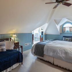 Attic bedroom with a white and blue color scheme featuring a bed, a daybed, a small window, a lamp, and a ceiling fan. An adjoining room is visible in the background.