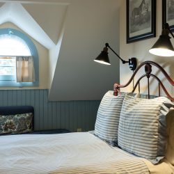 A cozy bedroom with a bed featuring striped bedding, two wall-mounted lamps, a blue cushioned window seat, and a half-moon window with a shade. Paintings hang on the wall above the bed.