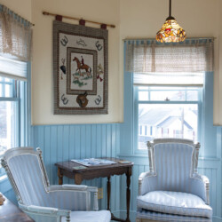 A cozy corner features two upholstered armchairs, a small wooden table, a hanging tapestry, a stained glass pendant light, and light blue paneling with windows offering outdoor views.