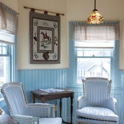 A cozy corner features two upholstered armchairs, a small wooden table, a hanging tapestry, a stained glass pendant light, and light blue paneling with windows offering outdoor views.