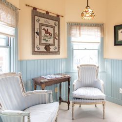 A small room with light blue wainscoting, two upholstered armchairs, a wooden side table, and horse-themed artwork on walls. Windows with sheer curtains allow natural light in.