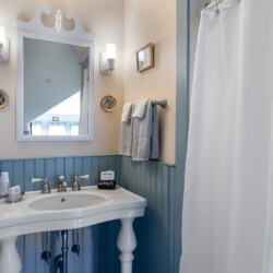 A small, well-lit bathroom with a white pedestal sink, wall-mounted mirror, light fixtures, towel rack, framed picture decor, and a shower curtain.