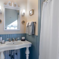 A small, well-lit bathroom with a white pedestal sink, wall-mounted mirror, light fixtures, towel rack, framed picture decor, and a shower curtain.