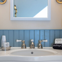 A bathroom sink with a round mirror above, white towels to the right, and a makeup bag labeled "makeup" on the counter. White and silver fixtures are seen on the sink.
