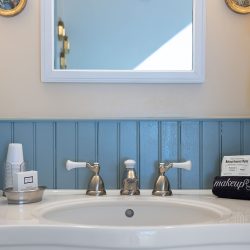 A bathroom sink with a round mirror above, white towels to the right, and a makeup bag labeled "makeup" on the counter. White and silver fixtures are seen on the sink.